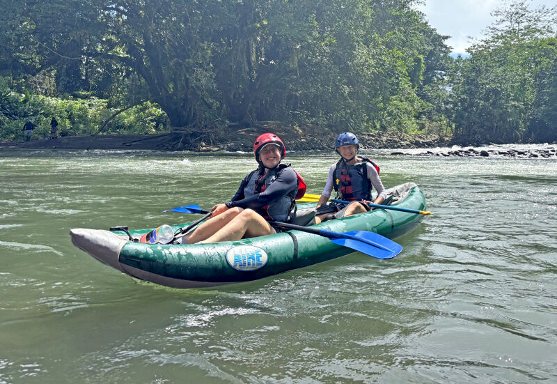 Two people are enjoying a sunny day while rafting on a river. They are in an inflatable raft, paddling through the water. The person in the front is wearing a red helmet and a black life jacket, while the person in the back has a blue helmet and a similar life jacket. Lush green trees line the riverbank, adding to the scenic beauty of the scene.